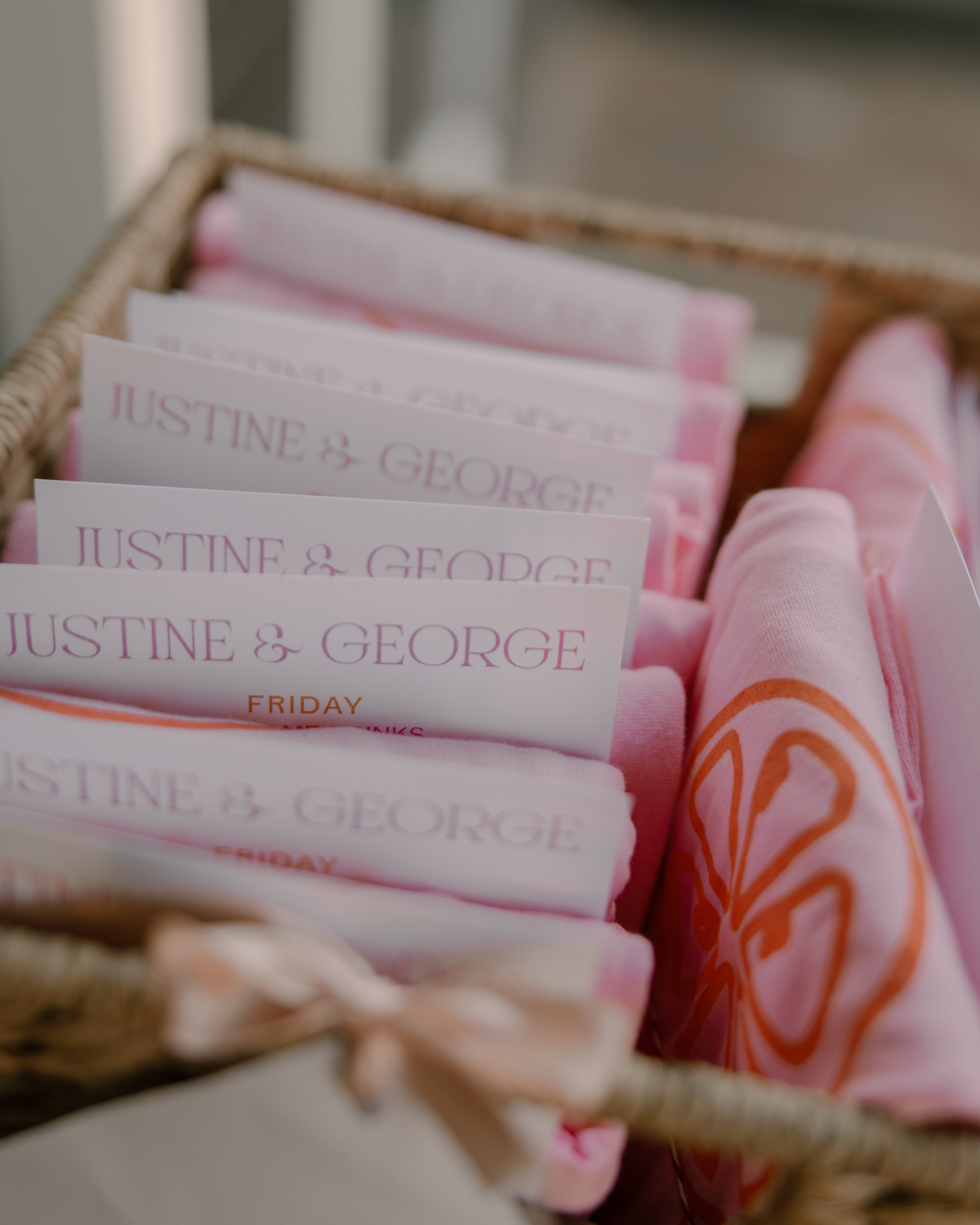 Stack of pink and white napkins with 'Justine & George' branded wedding itineraries in a woven basket.