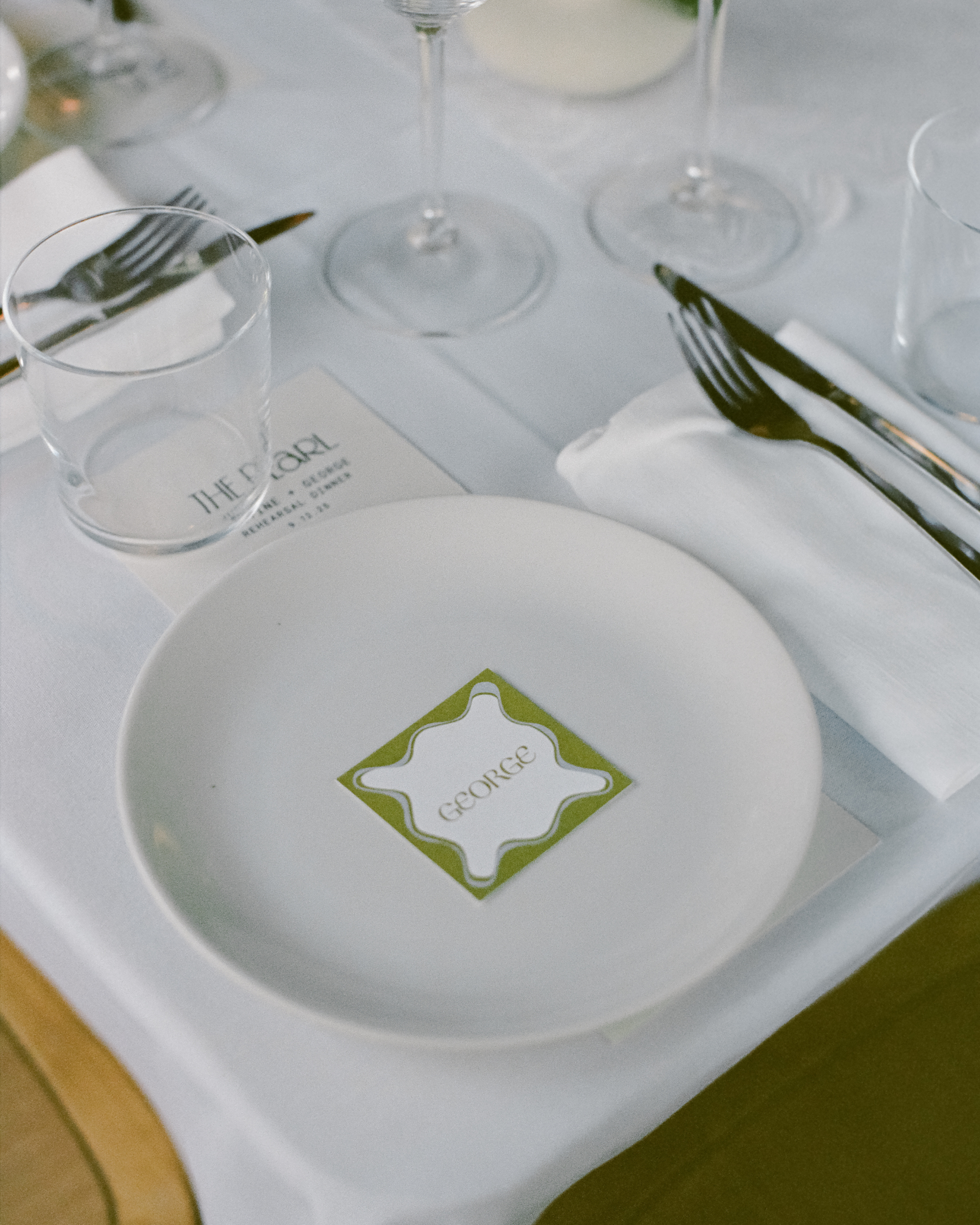 Dining table setting with a white plate, cutlery, custom design name tags and glasses on a white tablecloth.