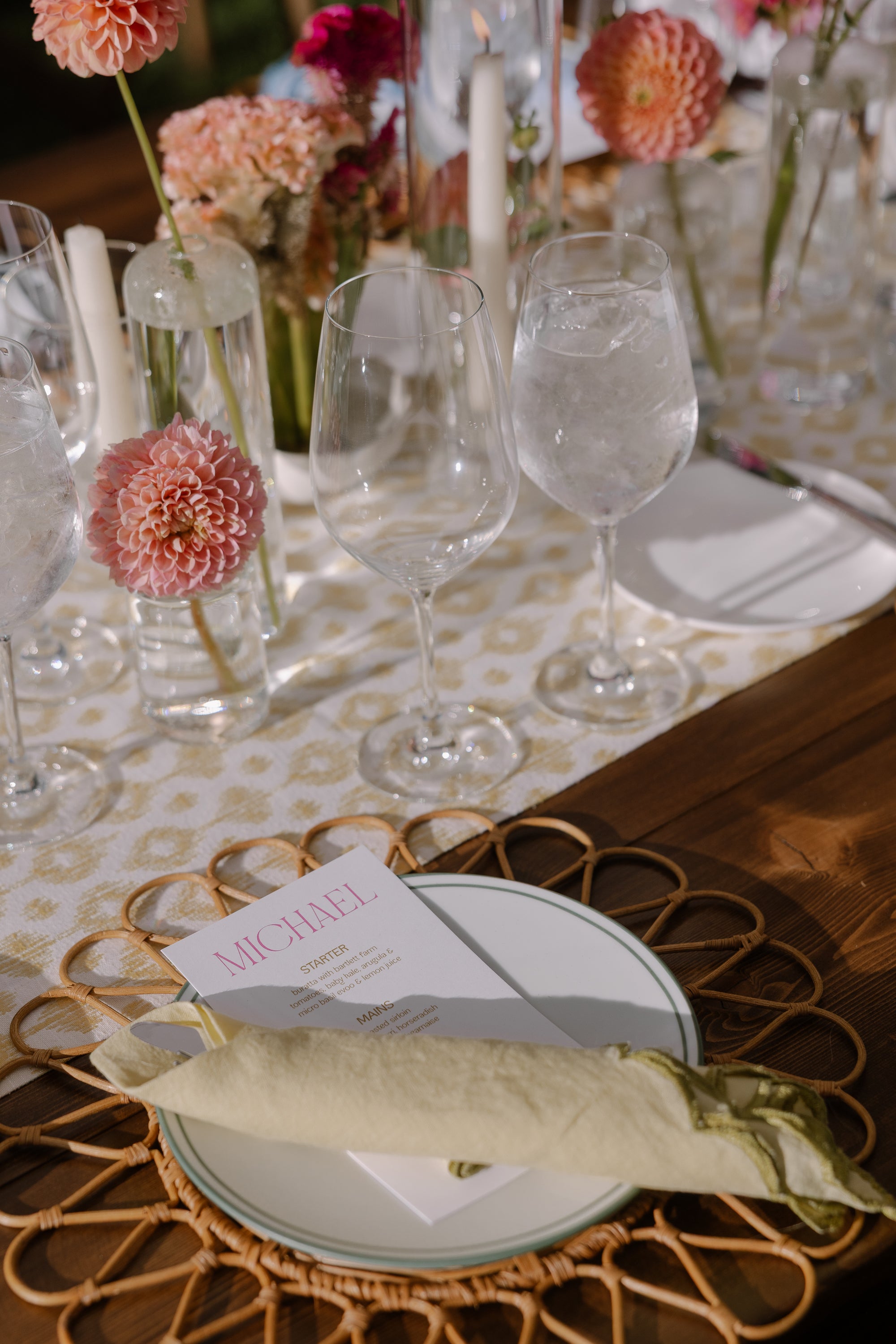 Table setting with wine glasses, flowers, and a place card on a patterned tablecloth.