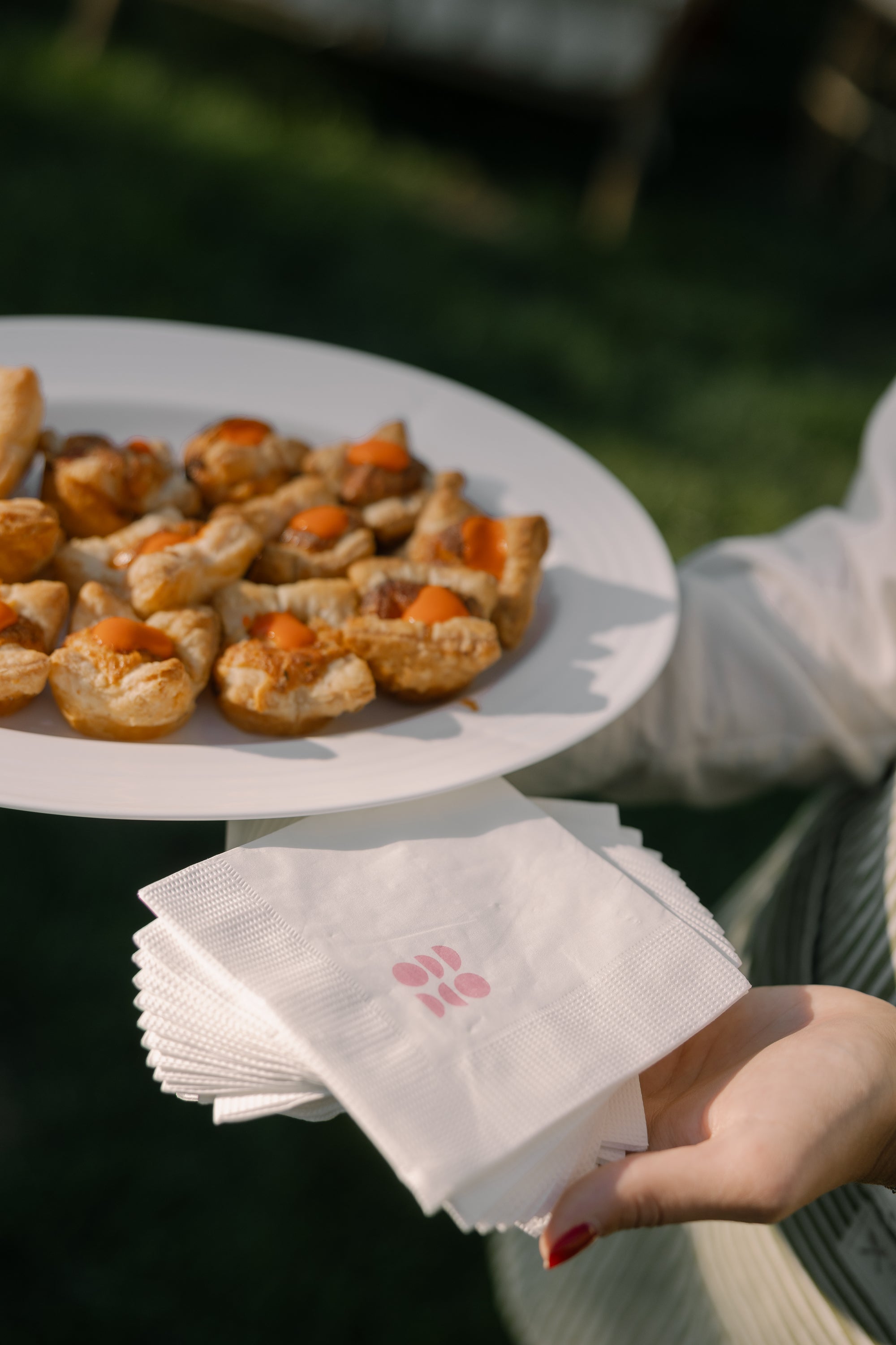 Person holding a stack of white napkins with a custom illustration, next to a plate of pastries outdoors.