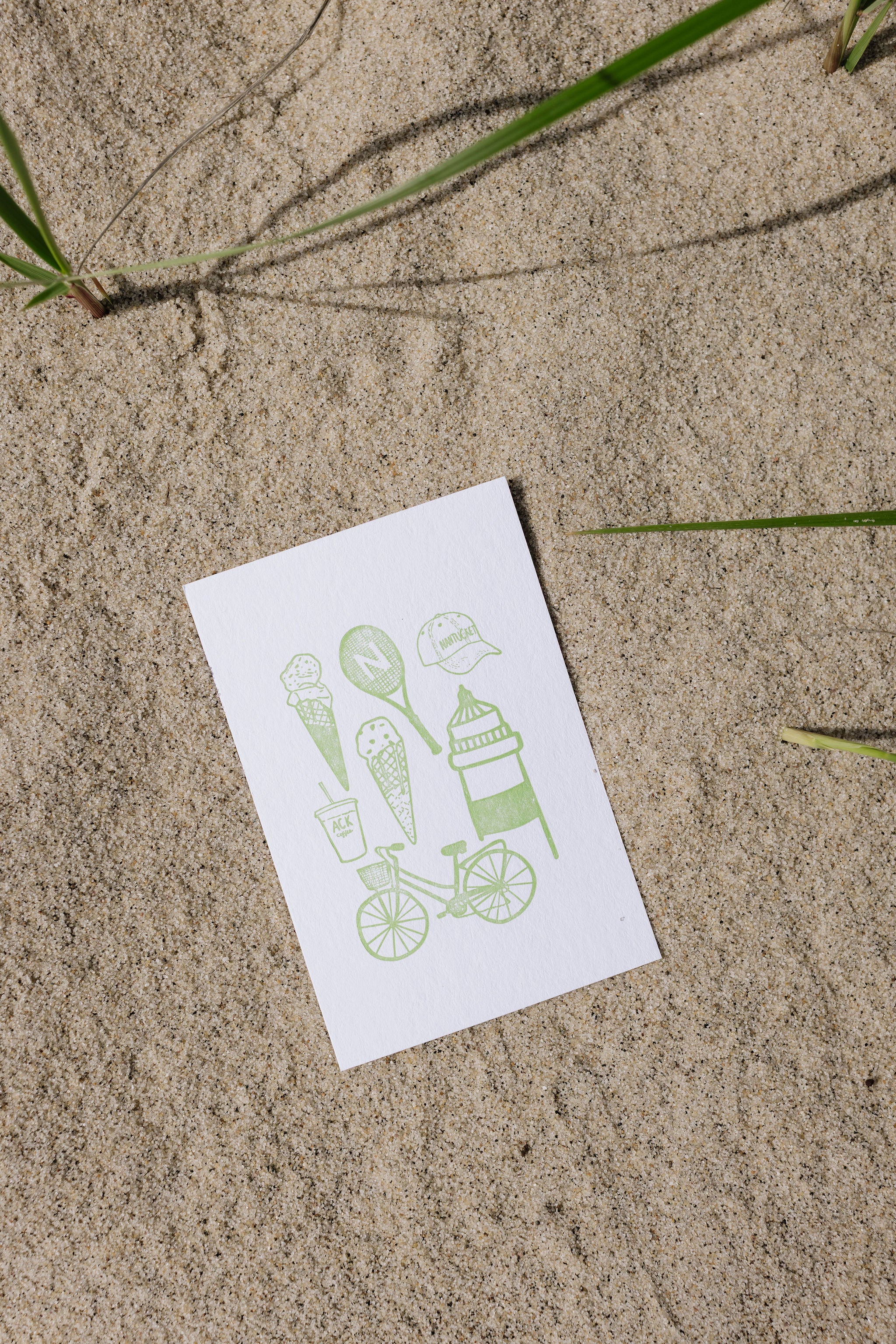 White postcard with green illustrations of ice cream cones, a bicycle, and other items on a sand surface.