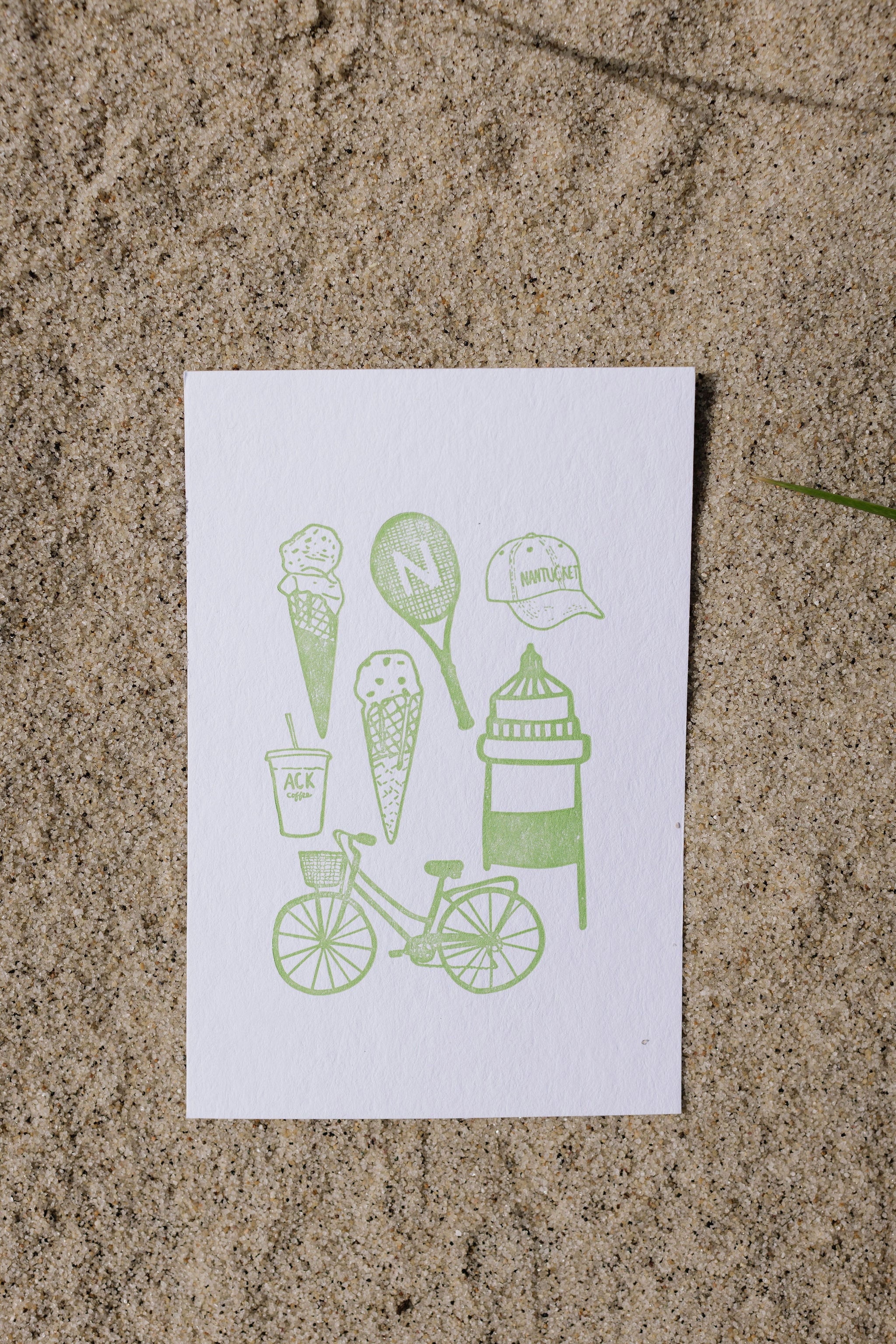 White postcard with green illustrations of ice cream cones, a bicycle, and other items on a sand surface.