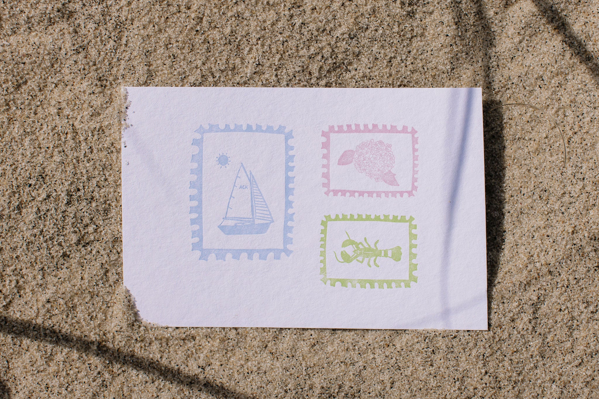 White paper with printed stamps of a sailboat, flower, and lobster on a sandy background
