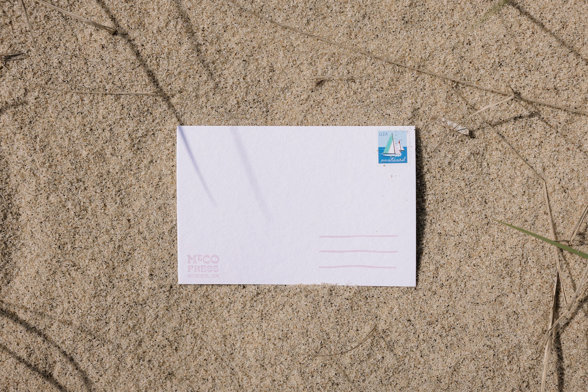 Postcard with pink writing lines and logo, with sailboat stamp on a sand surface.