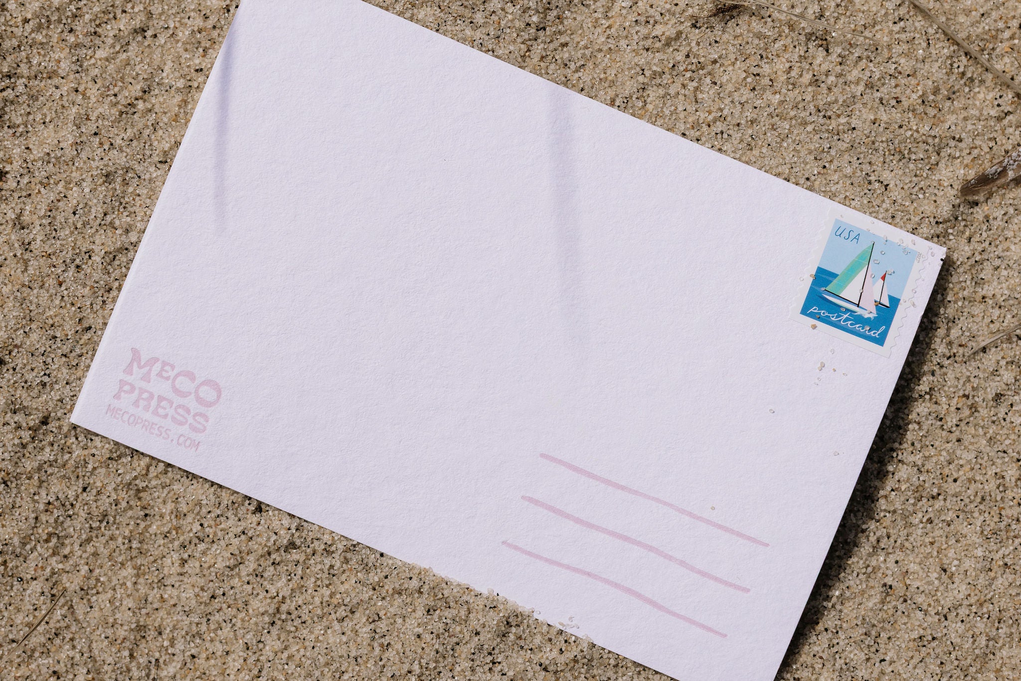 White postcard with a stamp and light link text and logo on a sand surface.