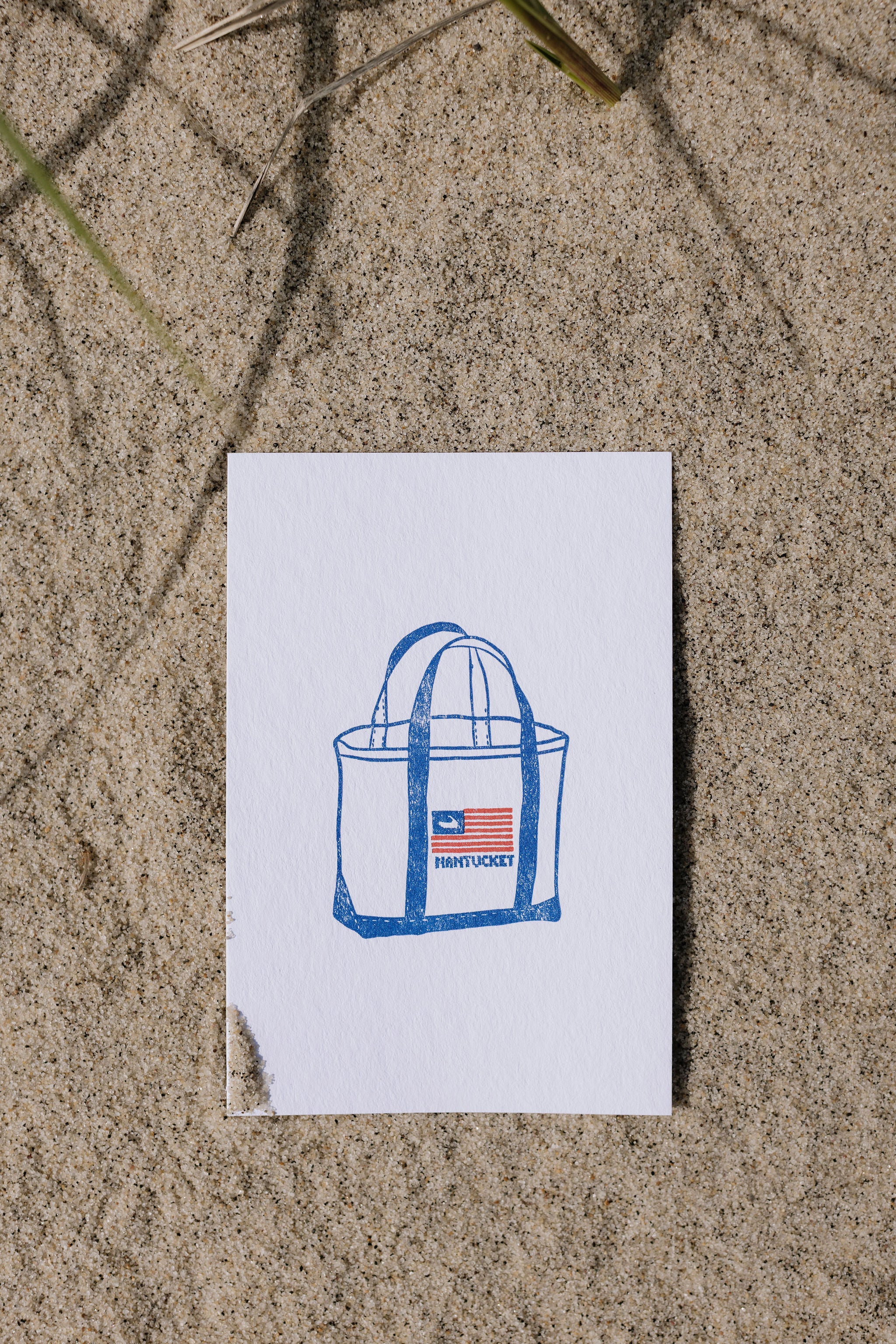 Postcard with a blue Nantucket flag tote bag illustration on a sandy surface
